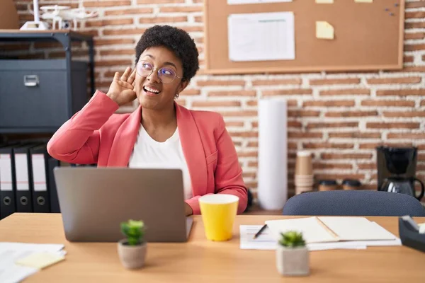 Young african american woman working at the office wearing glasses smiling with hand over ear listening and hearing to rumor or gossip. deafness concept.