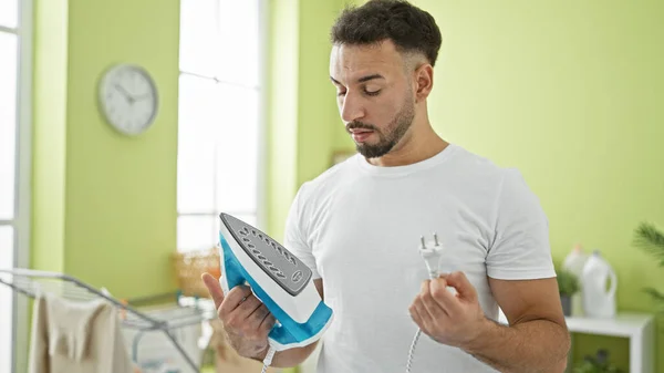 Young arab man holding ironing machine looking upset at laundry room