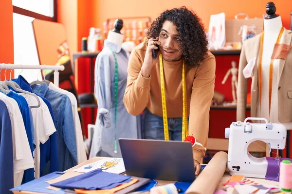 Genç Latin terzi atölyedeki laptopu kullanarak akıllı telefondan konuşuyor.