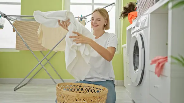 若い金髪の女性は 洗濯室で笑顔のバスケットに服を保持しています — ストック写真