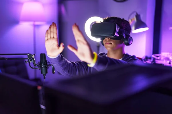 Young hispanic teenager streamer playing video game using virtual reality glasses at gaming room