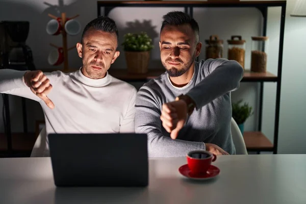Homosexual couple using computer laptop looking unhappy and angry showing rejection and negative with thumbs down gesture. bad expression.