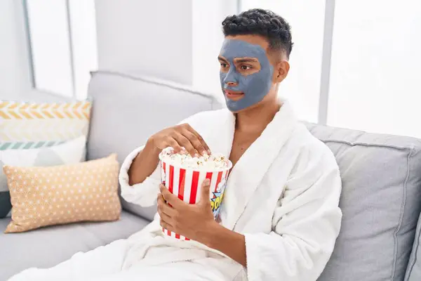 Young latin man relaxed on sofa with facial mask treatment eating popcorn at home