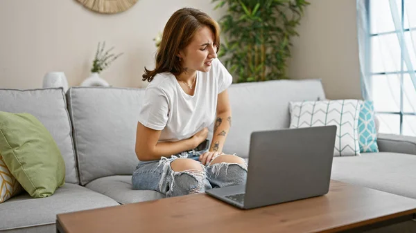 自宅で胃の痛みに苦しんでいるソファーに座っているノートパソコンを使用している若い女性 — ストック写真