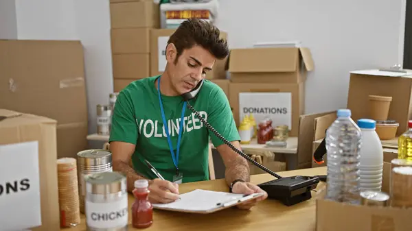 Young hispanic man volunteer talking on telephone writing on clipboard at charity center