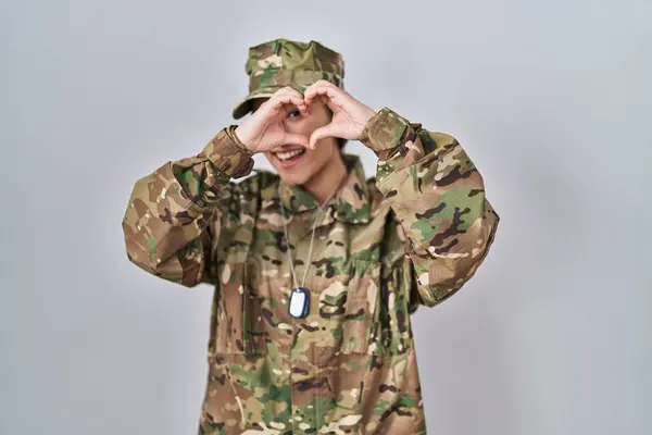 Young south asian woman wearing camouflage army uniform doing heart shape with hand and fingers smiling looking through sign