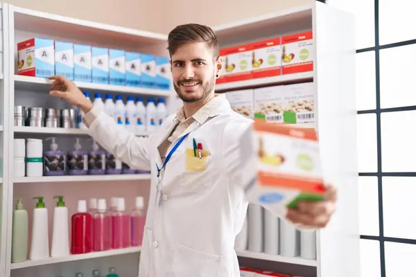 Joven Hombre Caucásico Farmacéutico Sonriendo Confiado Sosteniendo Paquete Vitaminas Farmacia —  Fotos de Stock