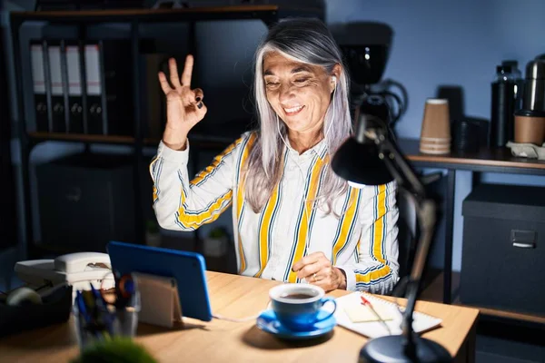 夜にオフィスで働く灰色の髪を持つ中年の女性は ポジティブに笑顔で手と指でOkサインします 成功した表現 — ストック写真