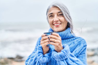 Middle age grey-haired woman smiling confident drinking coffee at seaside