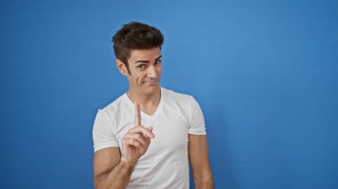 Genç İspanyol adam, izole edilmiş mavi arka planda gülümseyen parmağıyla hayır diyor.