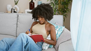 Evdeki koltukta oturan Afrikalı Amerikalı kadın deftere yazı yazıyor.
