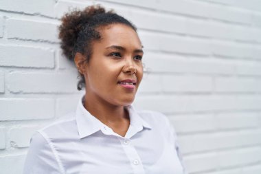 Beyaz tuğlalı arka planda ciddi bir ifadeyle yan tarafa bakan Afrikalı Amerikalı kadın.