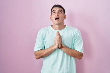 Yakışıklı İspanyol adam pembe arka planda duruyor. Elleriyle yalvarıyor ve dua ediyor. Yüzünde umut ifadesi var. Yalvarıyor. 