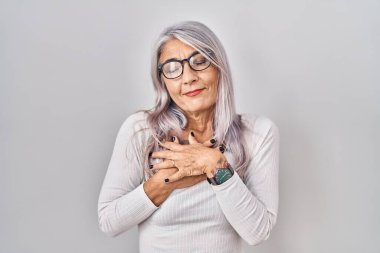 Beyaz arka planda beyaz saçlı, elleri göğsünde, gözleri kapalı ve yüzünde minnet dolu bir jest olan orta yaşlı bir kadın. sağlık kavramı. 
