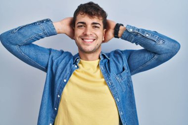 Young hispanic man standing over blue background relaxing and stretching, arms and hands behind head and neck smiling happy 