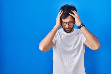 Mavi arka planda duran yakışıklı Latin adam acı ve migrenden dolayı çaresiz ve stresli baş ağrısı çekiyor. Eller başının üstünde.. 