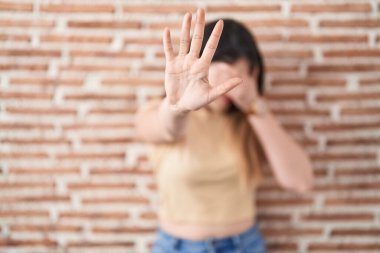 Genç esmer kadın tuğla duvarın üzerinde duruyor. Gözleri ellerle kaplıyor ve üzgün ve korku ifadeleriyle jestleri durduruyor. utangaç ve olumsuz kavram. 