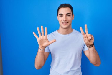 Young hispanic man standing over blue background showing and pointing up with fingers number seven while smiling confident and happy. 