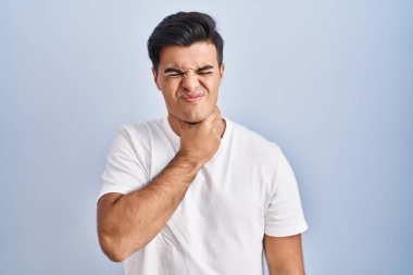 Mavi arka planda dikilen İspanyol adam ağrılı boynuna dokunuyor. Grip, mantar ve enfeksiyon yüzünden boğazı ağrıyor. 