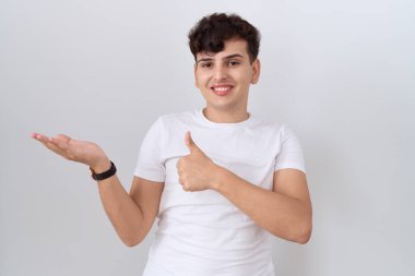 Gündelik beyaz tişört giyen genç bir adam avuç içini gösteriyor ve baş parmağıyla iyi bir hareket yapıyor, mutlu ve neşeli gülümsüyor. 