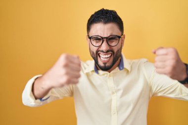 İspanyol genç adam iş kıyafetleri ve gözlük takıyor kızgın ve kızgın yumruklar sallıyor öfkeyle bağırırken sinirli ve öfkeli. öfke ve agresif konsept. 