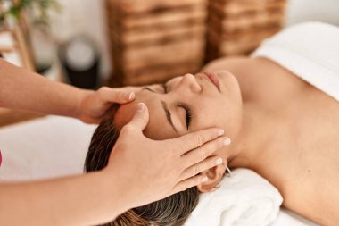 Young beautiful hispanic woman lying on table having head massage at beauty salon