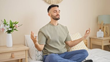 Young hispanic man doing yoga exercise sitting on bed at bedroom