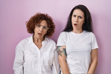 İspanyol anne ve kızı pembe arka planda günlük beyaz t-shirt giyiyorlar. Şüpheli ve alaycı görünüyorlar. Ağzını açınca şaşırıyorlar. 