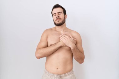 Beyaz arka plan üzerinde tişörtsüz duran genç İspanyol adam elleri göğsünde, gözleri kapalı ve yüzünde minnet dolu bir jest ile gülümsüyor. sağlık kavramı. 