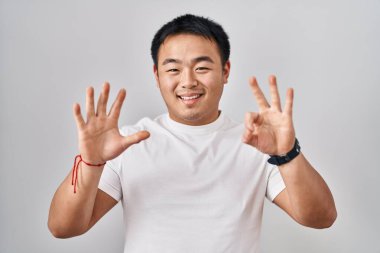 Beyaz arka planda duran genç Çinli adam sekiz numaralı parmaklarını gösterip işaret ederken kendinden emin ve mutlu gülümsüyor.. 