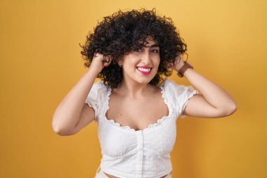 Sarı arka planda duran kıvırcık saçlı genç esmer kadın parmaklarıyla kulaklarını çekerek gülümsüyor, komik hareketler yapıyor. deneme sorunu 