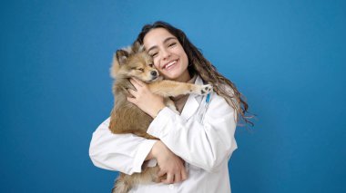 Köpek veterineriyle gülümseyen genç İspanyol kadın izole edilmiş mavi arka planda sarılıyor.