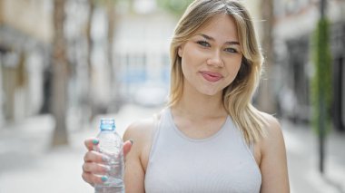 Genç sarışın kadın gülümsüyor. Elinde bir şişe su tutuyor.