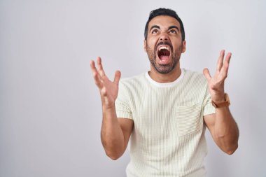 Hispanic man with beard standing over isolated background crazy and mad shouting and yelling with aggressive expression and arms raised. frustration concept. 
