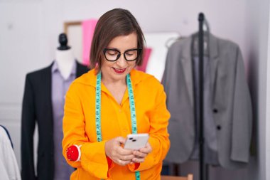 Middle age woman tailor smiling confident using smartphone at tailor shop