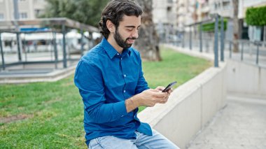 Genç İspanyol adam parkta gülümseyen akıllı telefon kullanıyor.