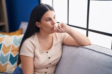 Young beautiful hispanic woman sitting on sofa with serious expression at home