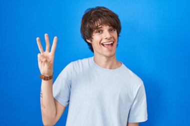 Hispanic young man standing over blue background showing and pointing up with fingers number three while smiling confident and happy. 