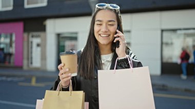 Güzel İspanyol bir kadın akıllı telefondan konuşuyor. Elinde alışveriş poşetleri ve sokakta kahve var.