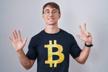 Beyaz, sarışın, bitcoin tişörtlü bir adam 8 numaralı parmaklarıyla onu gösterirken kendinden emin ve mutlu bir şekilde gülümsüyor.. 