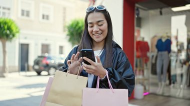 Genç, güzel, İspanyol bir kadın akıllı telefon kullanıyor. Elinde alışveriş torbaları var.