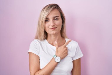Young blonde woman standing over pink background cheerful with a smile on face pointing with hand and finger up to the side with happy and natural expression 
