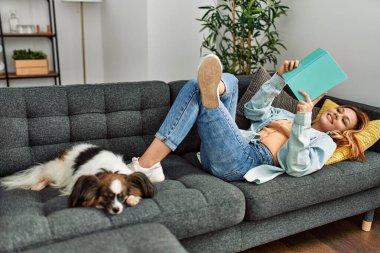 Genç beyaz kadın kitap okuyor. Evde köpekle birlikte kanepede yatıyor.