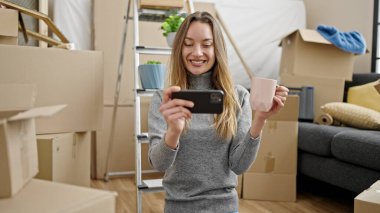 Beyaz bir kadın yerde oturmuş bir fincan kahve içiyor ve yeni evinde akıllı telefondan video izliyor.