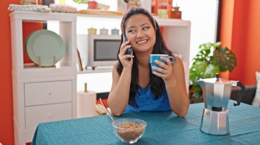 Genç Çinli kadın kahvaltı yapıyor. Yemek odasında akıllı telefondan konuşuyor.