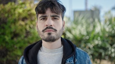 Young hispanic man standing with serious expression at park