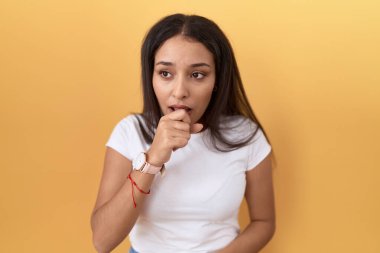 Sarı arka plan üzerine günlük beyaz tişört giyen genç Arap kadın kendini iyi hissetmiyor ve soğuk algınlığı veya bronşitin belirtisi olarak öksürüyor. sağlık hizmeti kavramı. 