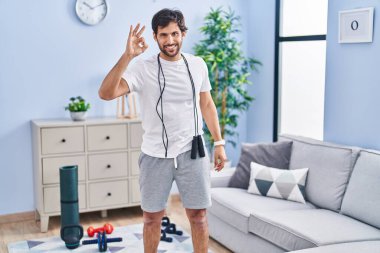 Evde spor kıyafetleri giyen yakışıklı Latin adam gülümsüyor ve el ve parmaklarıyla 