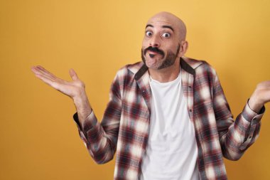 Sarı arka planda dikilen sakallı, kafası karışmış ve kolları ve elleri havada İspanyol bir adam. şüphe kavramı. 