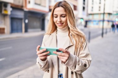Genç sarışın kadın gülümsüyor. Sokakta akıllı telefondan videolar izliyor.